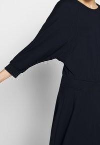 Czarna sukienka z luźnymi, szerokimi rękawami, żeberkowym pasem, gładką teksturą i minimalnym detalowaniem szwów. Odpowiednia do swobodnego noszenia.