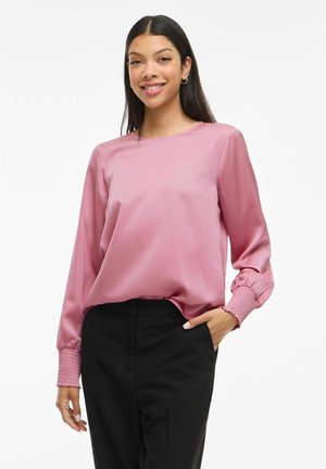 Femme souriante, portant un blouse en satin rose à manches longues avec des poignets smockés et un pantalon noir, main dans la poche, sur fond blanc.