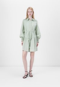 Hellgrünes Hemdkleid mit gerafften Ärmeln, Spitzenverzierung und Kordelzug in der Taille. Getragen mit schwarzen Sandalen mit Riemchen.
