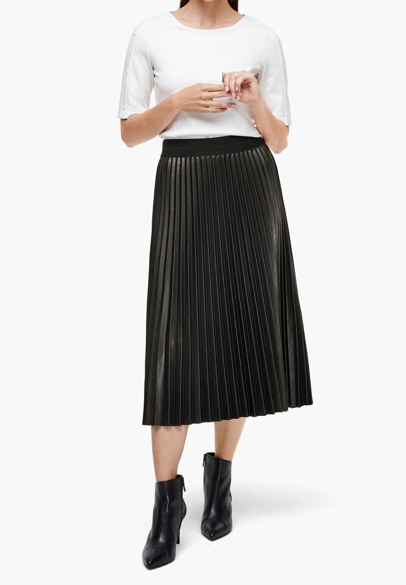 Falda midi negra con pliegues y un acabado texturizado, acompañada de una blusa blanca con detalles de botones en las mangas y botas negras hasta el tobillo.