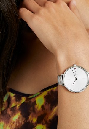 Poignet avec une montre en maille argentée minimaliste affichant 10 h 10, portée par une personne vêtue d'un haut coloré tie-dye, main près du visage avec des cheveux foncés.