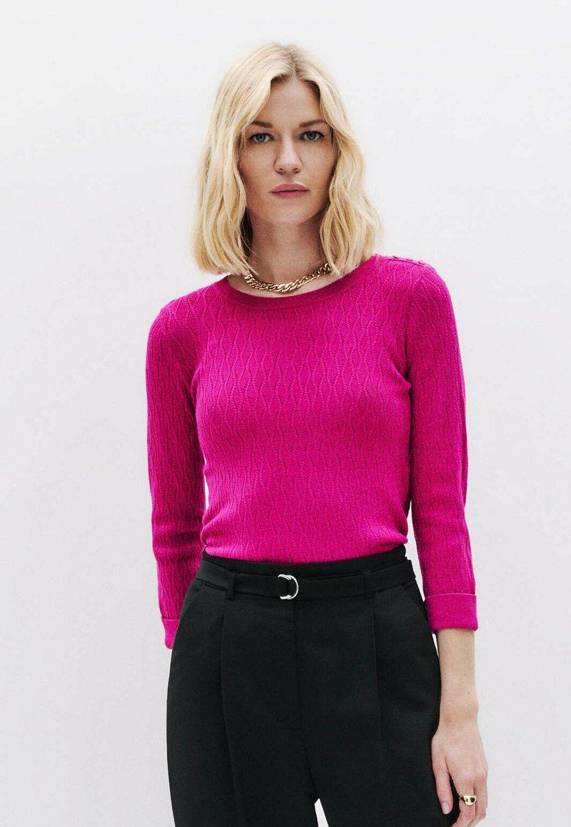 Fuchsia Strickpullover mit einem strukturierten Rautenmuster, Rundhalsausschnitt und Dreiviertelärmeln, kombiniert mit hochgeschnittenen schwarzen Hosen.