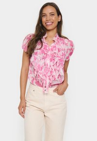 Rožinė gėlių bluzon�ėlė su raukiniais ir juosta ties juosmeniu; permatomas audinys su trumpomis pūstomis rankovėmis; dėvima su šviesiomis kelnėmis.