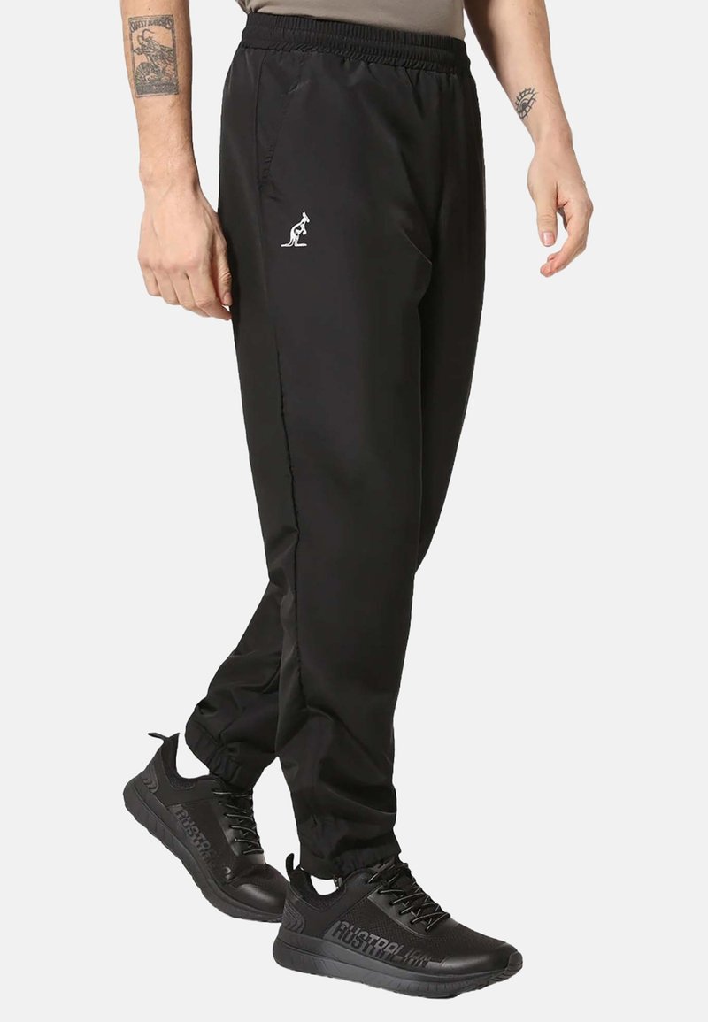 Pantaloni sportivi neri con elastico in vita, tasche laterali e un piccolo logo bianco. Indossati con scarpe da ginnastica nere dalla superficie testurizzata.