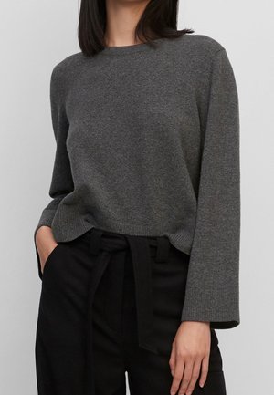 Kvinde iført en mørkegrå langærmet sweater stoppet ned i sorte bukser med høj talje og et bundet stofbælte.