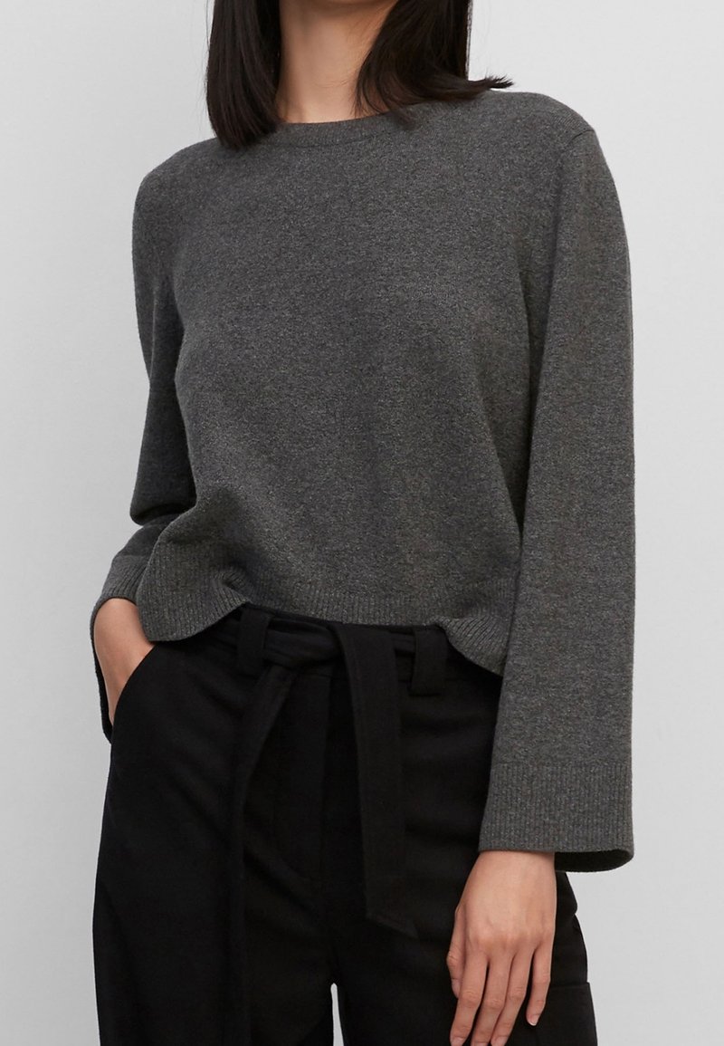 Femme portant un pull gris foncé à manches longues rentré dans un pantalon noir taille haute avec une ceinture en tissu nouée.