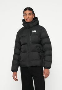 Homme portant une doudoune noire avec capuche et un logo "HH" blanc, se tenant contre un fond blanc uni, les mains sur les côtés.
