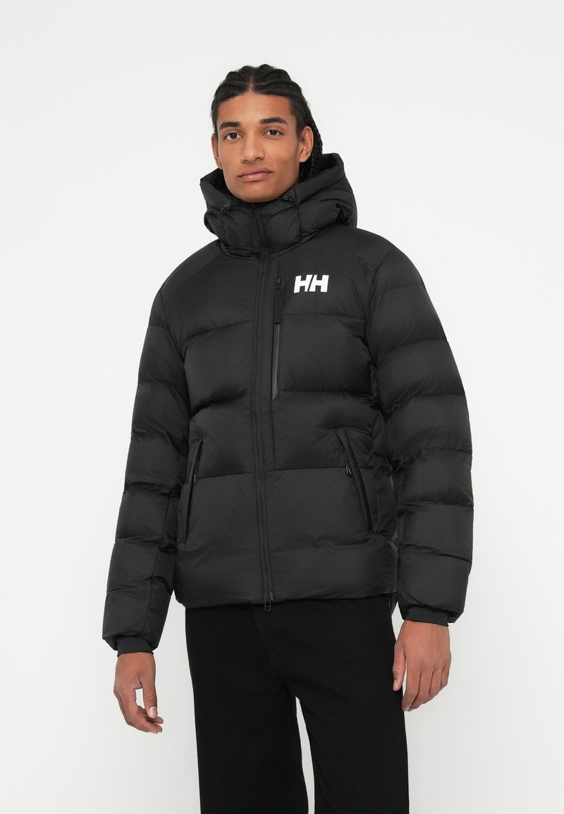 Homme portant une doudoune noire avec capuche et un logo "HH" blanc, se tenant contre un fond blanc uni, les mains sur les côtés.