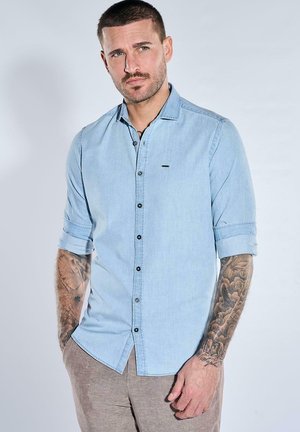 Mann mit kurzen braunen Haaren und Bart, trägt ein hellblaues hochgekrempeltes Hemd und beige Hosen, zeigt detaillierte Tätowierungen auf beiden Unterarmen.