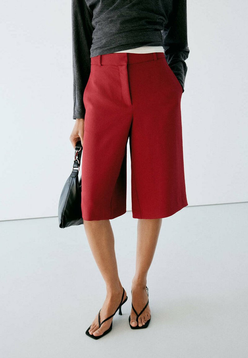 Personne portant un short rouge sur mesure arrivant aux genoux, des sandales à talons noires, un haut sombre à manches longues, tenant un petit sac à main noir dans une main.