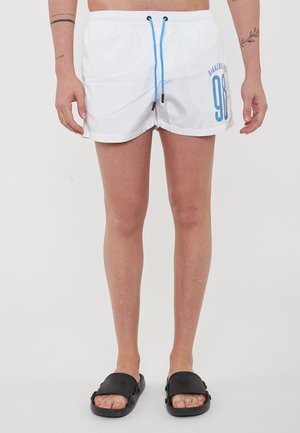 Weiße Schwimmshorts mit einem blauen Kordelzug, auf der linken Seite mit einer Grafik "98". Getragen mit schwarzen Slidesandalen. Aus glattem, leichtem Material.