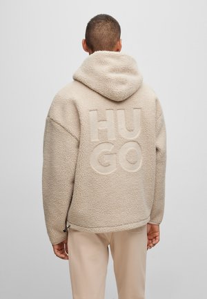 Osoba ubrana w beżową polarową kurtkę z kapturem z dużymi, wypukłymi literami „HUGO” na plecach, w połączeniu z jasnymi spodniami.