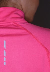 Nahaufnahme des oberen Rückens und Nackens einer Person, die ein leuchtend pinkes Sportshirt mit sichtbaren Nähten und reflektierenden Streifen trägt.