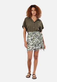 Camicia corta verde oliva con colletto, abbinata a una gonna bianca con motivi di foglie di palma verdi e dettagli in frange. Sandali neri.