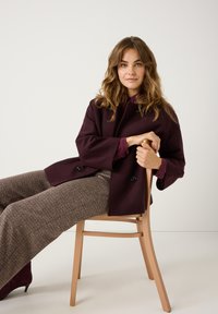 Bordeauxjas met brede mouwen over een paarse blouse, gecombineerd met gestreepte, brede broek. Model zit op een lichthouten stoel.
