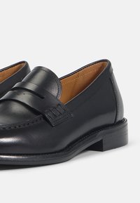Mocassins penny en cuir noir avec talons bas, détails cousus et doublure intérieure couleur tabac.