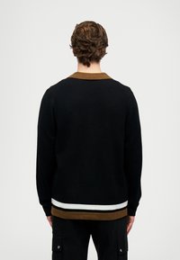 Férfi hátulról, fekete pulóvert visel barnával díszített gallérral és aljjal, fehér és fekete csíkokkal az alján, valamint fekete cargo nadrággal.