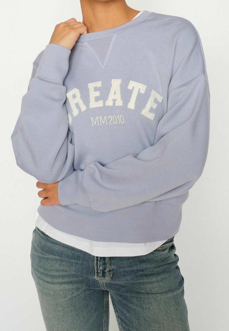Personne portant un sweat-shirt violet clair avec les mots "CREATE" et "MM 2010" en lettres crème, associé à un jean bleu et une chemise blanche.