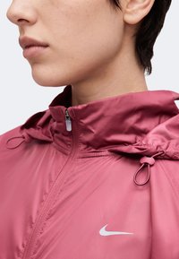 Κοντινό πλάνο ατόμου που φοράει ροζ ζακέτα Nike με φερμουάρ και ρυθμιζόμενα κορδόνια κουκούλας, εμφανίζοντας το κάτω μέρος του προσώπου και τον λαιμό.