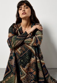  Lange jurk met een patroon van een mix van donkergroene, bruine en gouden tinten, met geometrische en bloemmotieven, en met pofmouwen.