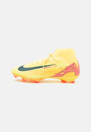 Chaussure de football jaune avec un col chaussette, présentant un swoosh vert foncé, des accents roses pixélisés et des crampons rouges sur une surface texturée.