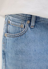 Close-up af lyseblå denimjeanslomme med metalnitter og orange syninger, båret med en hvid top.