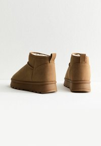 Botas de tobillo de ante marrón con un forro suave, punta redondeada y suelas de goma que presentan un patrón de agarre texturizado; incluyen una lengüeta en la parte trasera.