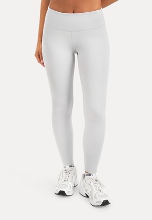 Kvinde iført hvide leggings med taljehøjde og hvide sportssko, stående på en ensartet lys baggrund.