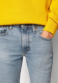 Pantalones vaqueros de mezclilla azul claro con un acabado ligeramente desgastado, que cuentan con bolsillos delanteros, costuras naranjas y una textura sutil. Combinados con una sudadera amarilla.