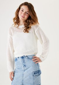 Un pull blanc en tricot avec un motif en maille ouverte, col rond et manches longues, associé à une jupe en denim bleu clair dotée de poches.