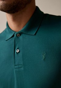 Polo verde con un tejido texturizado, dos botones en el cuello y un logo de ciervo bordado en el pecho izquierdo.