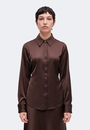 Personne aux cheveux courts et foncés porte une chemise marron en satin à manches longues boutonnée, debout devant un fond clair uni.