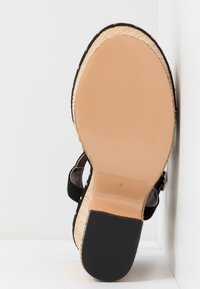 Sandale compensée noire avec une semelle en espadrille texturée, un bout rond, une bride de cheville ajustable avec accents métalliques, et une semelle intérieure beige lisse.