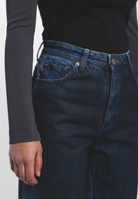 Jeans de mezclilla oscuros de cintura alta con corte recto. Incluyen cinco bolsillos, una bragueta con botones y detalles de costura sutiles.