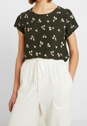 Kobieta ubrana w ciemnozieloną bluzkę z krótkim rękawem z beżowym nadrukiem wiśni oraz białe, wysokie spodnie z paskiem ściągającym i pionowymi paskami.