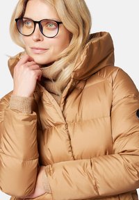  ocieplana kurtka z kapturem w jasnym brązie, z połyskującym wykończeniem, prążkowanymi mankietami i bocznym akcentem z logo. Pod nią miękki sweter z dzianiny.