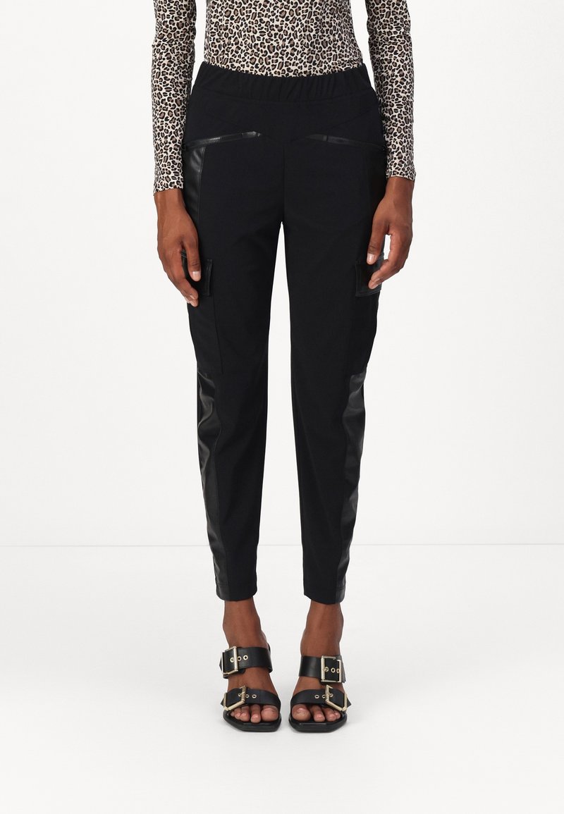 Femme portant un pantalon noir fuselé avec fermeture éclair et poches plaquées, associé à un haut à manches longues imprimé léopard et des sandales noires à boucles.