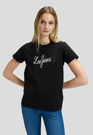 Giovane donna in piedi con le mani sui fianchi, indossa una t-shirt nera "Lee Jeans" e jeans blu su uno sfondo grigio chiaro e semplice.
