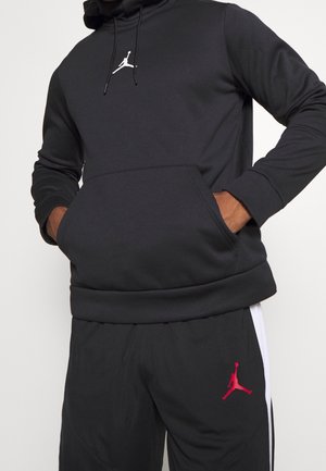 Person trägt schwarzen Kapuzenpullover und schwarze Hose mit weißen und roten Jumpman-Logos, Hände in der vorderen Tasche des Kapuzenpullovers.