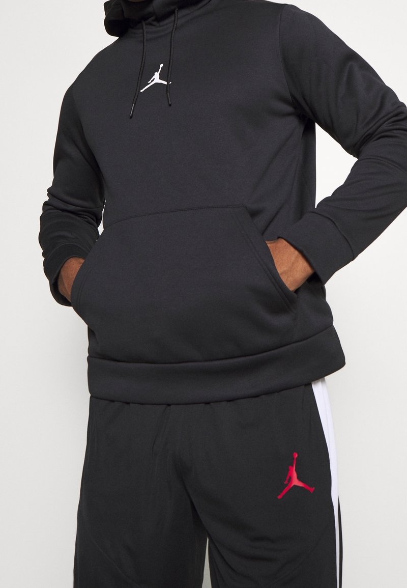 Personne portant un sweat à capuche noir et un pantalon noir avec des logos Jumpman blancs et rouges, mains dans la poche avant du sweat.