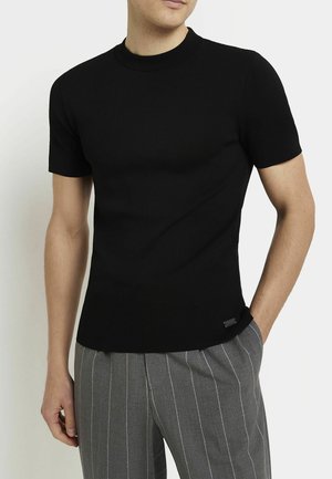 Mężczyzna ubrany w dopasowaną czarną koszulkę z krótkim rękawem i szare spodnie w cienkie paski, stojący z jedną ręką w kieszeni na tle białego tła.