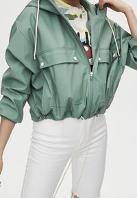 Chaqueta de cremallera verde claro con capucha, dos bolsillos frontales, dobladillo elástico y cordón. Combinada con una camisa estampada y pantalones cortos blancos.