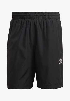Man draagt een donkergroen Adidas T-shirt, zwarte Adidas shorts en witte Adidas sneakers met zwarte strepen, staand met handen in zakken.