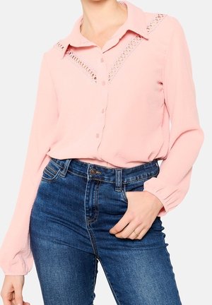 Vrouw draagt een lichtroze blouse met kanten uitsnijdingen op de schouders en blauwe jeans met hoge taille, hand rustend nabij de taille.