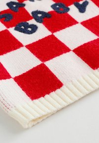 Červeno-bílý kostkovaný pletený materiál s žebrovaným krémovým okrajem a tmavě modrými kurzívními písmeny vyšitými blízko vrchu.