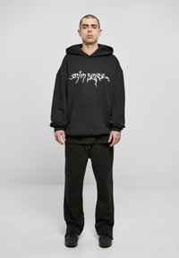Schwarzer oversized Hoodie mit einem weißen Grafikdruck und einer Fronttasche, kombiniert mit passenden schwarzen Jogginghosen und schwarzen Schuhen. Schlichtes, lässiges Design.