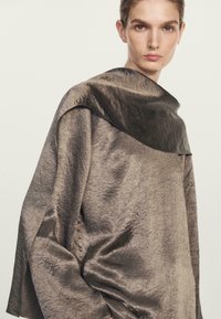 Metaliczny brązowy top z drapowanym, wysokim kołnierzem. Posiada teksturowane wykończenie oraz szerokie rękawy, o luźnym i swobodnym kroju.