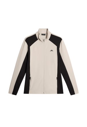 Lys beige zip-jakke med sorte sidepaneler, høj krave, lange ærmer og logo på brystet. Glat, let materiale.