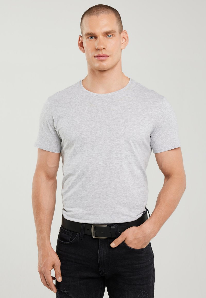 Ung mand med kort hår iført en tætsiddende lysegrå t-shirt, sort bælte og sorte jeans, stående mod en ensfarvet lys baggrund.