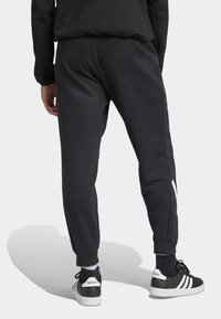 Person som bär svarta avsmalnande mjukisbyxor med vitt logotyp och svarta Adidas-sneakers med vita ränder, sedd bakifrån mot en vit bakgrund.